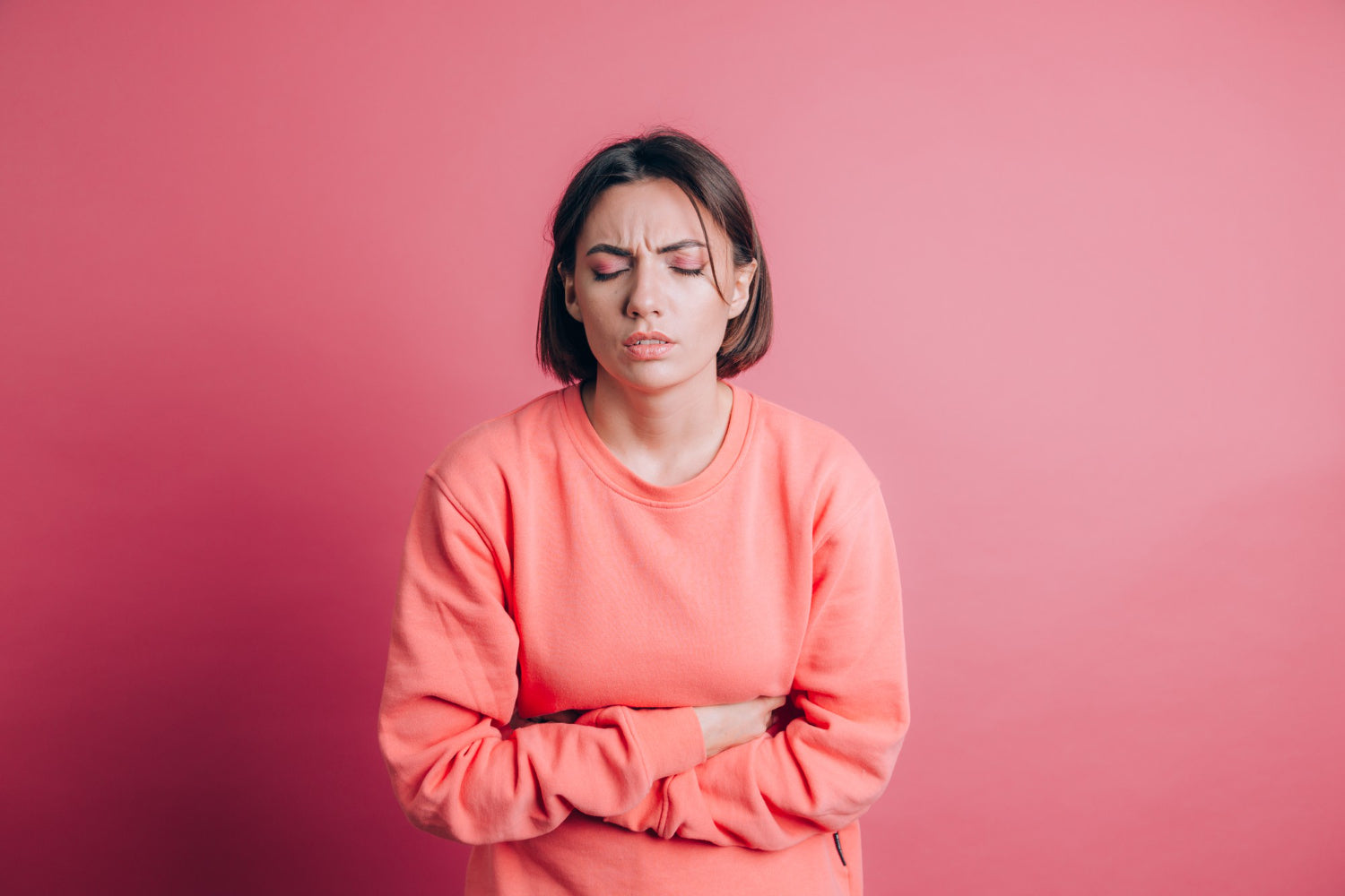 Image of a woman wearing a peach sweater against a pink wall suffering from stomach ache and constipation with painful grimace. 