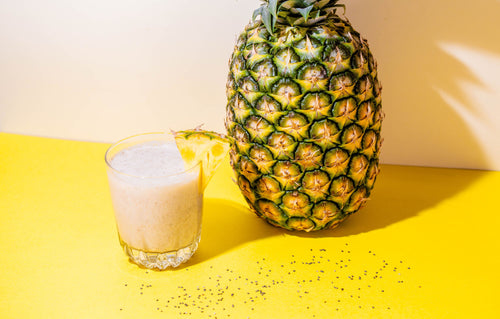 a glass of Pineapple Chia Cleanse beside a pineapple on a yellow surface against a white wall. 