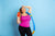 Image of a curvy woman holding an orange water jug on her right hand, her left hand on her forehead, and a hula hoop on her left shoulder. She is standing against a blue wall.