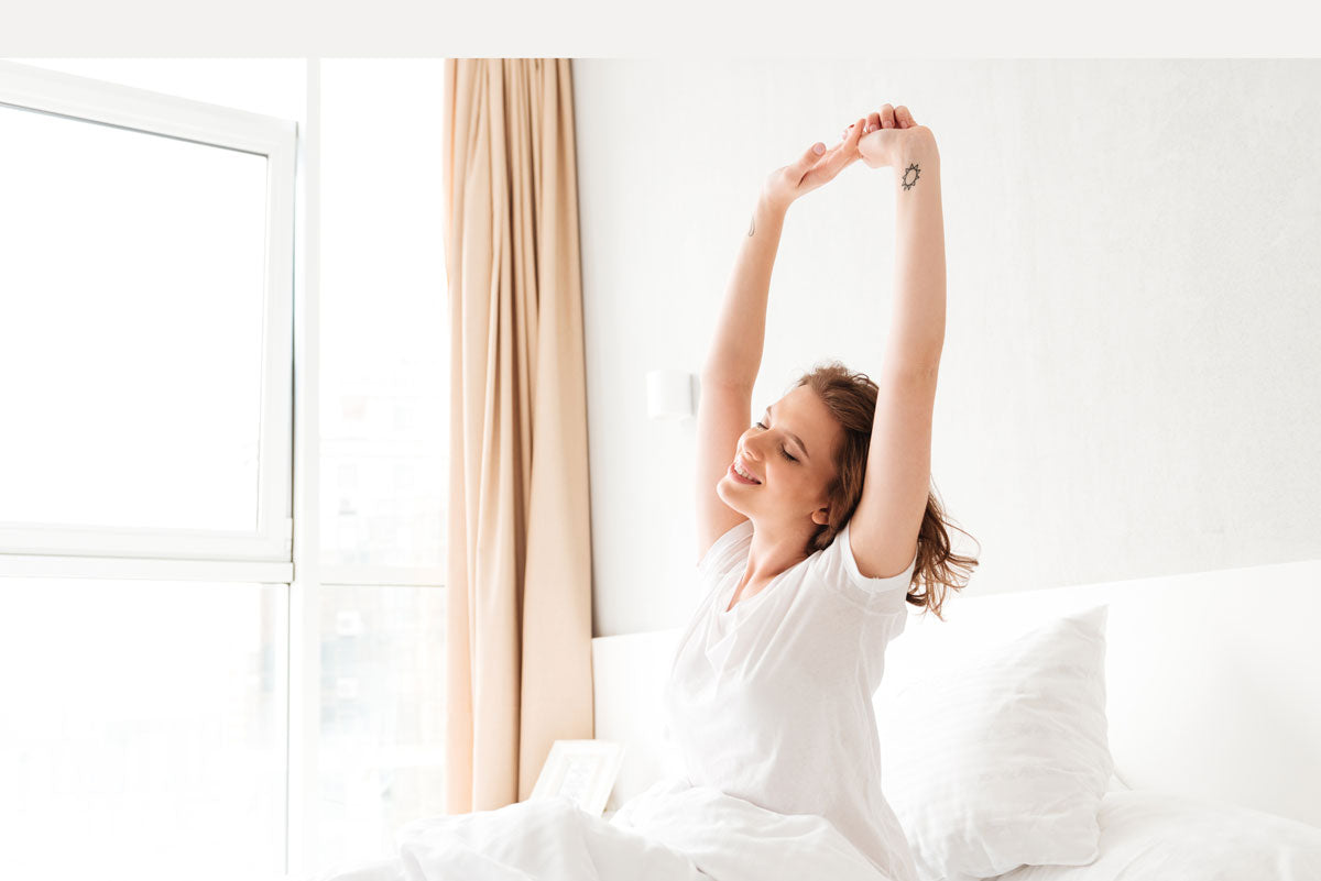 Image of a woman who just woke up, still sitting on her bed, and stretching her arms in an upward pose.