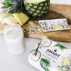 A sliced-in-half pineapple and in front is 3 slices of pineapple, some light brown powder and a single serving sachet of Pineapple Chia Cleanse on a wooden board. On the white surface is a glass of white smoothie beside a box of Smart Pressed Juice package.