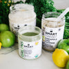 One 500 grams open jar of Vegan Vanilla Proteini, in front is one 240 grams open jar of Organic Pressed Greens beside one 375 grams open jar of Pineapple Chia Cleanse surrounded by green vegetables, 1 lemon, and green apples.