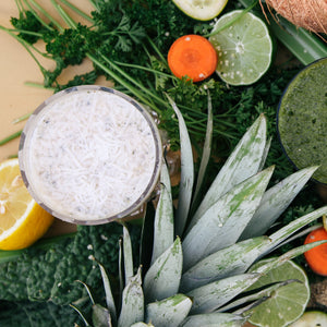 A glass of white smoothie surrounded by different vegetables like parsley, slices of carrots and some slices of lemon and lime, and a pineapple on a wooden surface.