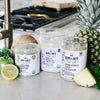One 240 grams open jar of Organic Pressed Greens, one 500 grams open jar of Vegan Vanilla Proteini, and one 375 grams  open jar of Pineapple Cleanse side by side surrounded by a slice of pineapple, parsley, whole pineapple, 1 coconut, 1 apple, and half an orange in a kitchen setting.