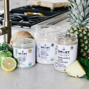 One 240 grams open jar of Organic Pressed Greens, one 500 grams open jar of Vegan Vanilla Proteini, and one 375 grams  open jar of Pineapple Cleanse side by side surrounded by a slice of pineapple, parsley, whole pineapple, 1 coconut, 1 apple, and half an orange in a kitchen setting. 