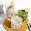 1 tall glass of white smoothie with chia seed toppings, a straw, and a slice of pineapple, a small glass of white smoothie with green garnish, and a small glass of green smoothie with a slice of lemon. The 3 glasses are all placed in one wooden coaster.