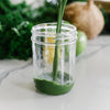 A glass being poured with a green smoothie. In the background is a blurred green vegetables, coconut, lemons, and limes.