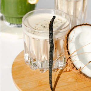 A glass white smoothie with a stick of vanilla standing against it and beside is an open coconut, both is on a wooden coaster. Behind is a glass of green smoothie beside a glass of white smoothie on a white surface.