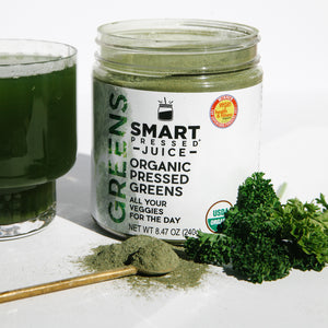 One 240 grams open jar of Organic Pressed Greens beside a glass of green smoothie and green herbs on the other side, and a stirring spoon full of navy green powder on the surface against a white background.