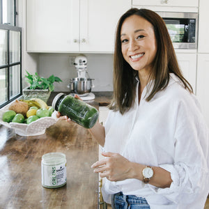A beautiful and healthy-looking woman holding a jar of green smoothie. On the kitchen top is an open jar of 240 grams Organic Pressed Greens and fruit basket full of different fruits. 