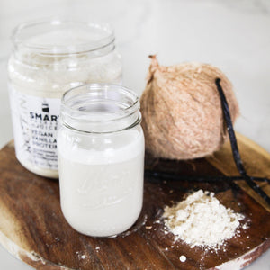 One 500 grams open  jar of Vegan Vanilla Proteini beside a coconut with 2 sticks of vanilla standing against it in the back row. In the front row is a jar of white liquid and light brown powder all are on a circular wood coaster. Set against a white background.