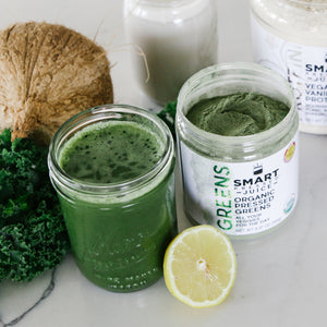 A glass of green smoothie beside an open jar of 240 grams of Organic Pressed Greens and a slice of lemon in the middle of those jars. Behind is some parsley, coconut, a glass of white smoothie, and a jar of Vegan Vanilla Proteini on a white surface.