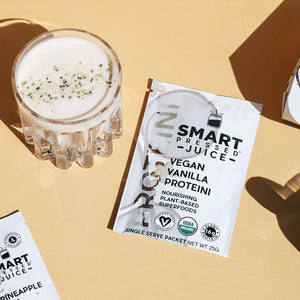 A glass of white smoothie with chia seed topping beside a single-serving packet of Vegan Vanilla Proteini on a beige surface.