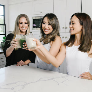 3 beautiful and healthy-looking ladies each is holding a glass of Smart Pressed Juice in different flavors and making a toast. The setting is in a kitchen top.