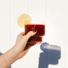 A hand of a woman holding a glass of red smoothie with a slice of lemon against a white wall with a shadow of her hand.