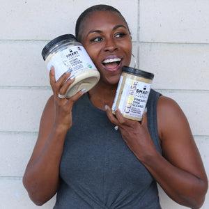 Beautiful black woman holding one Vegan Vanilla Proteini jar in one hand and one Pineapple Chia Cleanse jar in the other hand. She is standing against a brick wall.