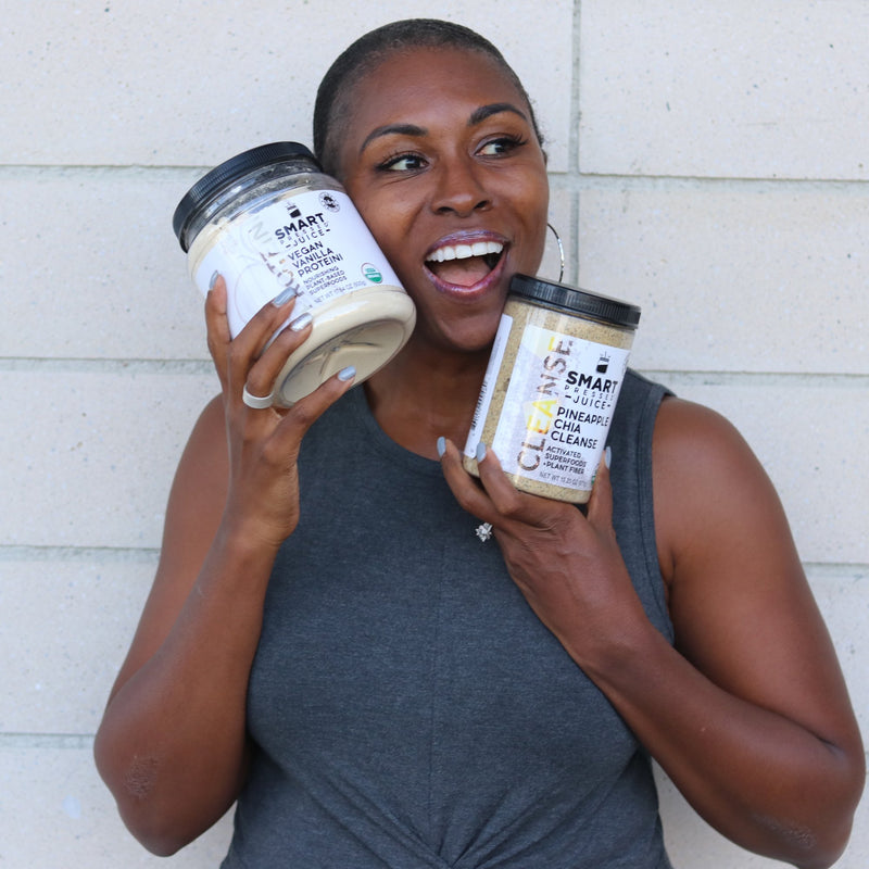Beautiful black woman holding one Vegan Vanilla Proteini jar in one hand and one Pineapple Chia Cleanse jar in the other hand. She is standing against a brick wall.