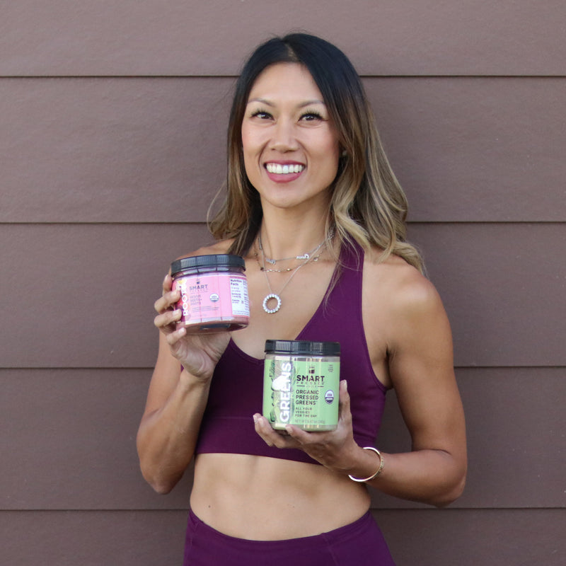 A beautiful and healthy-looking woman holding a jar of Revive Beet+Roots on the right and a jar of Organic Pressed Greens on the left. She is standing against a brown wall.