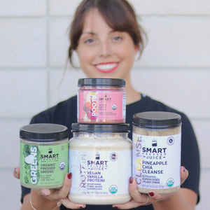 A woman holding 1 jar of 150 grams Revive Beet + Roots on the top and 1 jar of 240 grams Organic Pressed Greens, 1 jar of 500 grams Vegan Vanilla Proteini, and 1 jar of 375 grams Pineapple Chia Cleanse side by side at the bottom row. She Is standing against a brick wall.