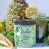 1 jar of 240 grams Organic Pressed Greens beside a jar of green smoothie with a slice of orange, beside is a pineapple fruit, broccoli, carrots, and lemon side by side against a light blue background.