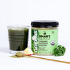 A glass of green smoothie beside a jar of 240 grams Organic Pressed Greens and a parsley next to it. In front is a slice of apple and a stirring spoon full of green powder. Set against a white background.