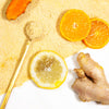 Light brown powder on the surface with a slice of turmeric on the top, 2 slices of oranges on the right, a stirring spoon full of powder, a slice of lemon, and a slice of ginger on a white surface.