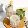 Three glasses of smoothies - one smoothie in a tall glass containing a beige colored smoothie with chia seeds on top and a pineapple wedge. The smaller glass has a greens smoothie and a lemon wedge.  The smaller glass that is in front has a beige colored smoothie with seeds on top. Image set against white background. glasses are on a small wooden lazy susan.