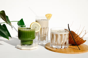 Green leaves beside a glass of green smoothie with a slice of lemon in a white coaster, a tall glass of white smoothie with pineapple wedge and a straw, a glass of white smoothie with a stick of vanilla standing against it, and half a coconut in a wooden coaster. Set against a white background.